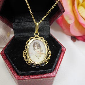 May include: A gold-toned pendant necklace featuring an oval portrait of a woman with a decorative filigree frame. The pendant hangs from a delicate chain and is displayed in a red jewelry box with a black interior.