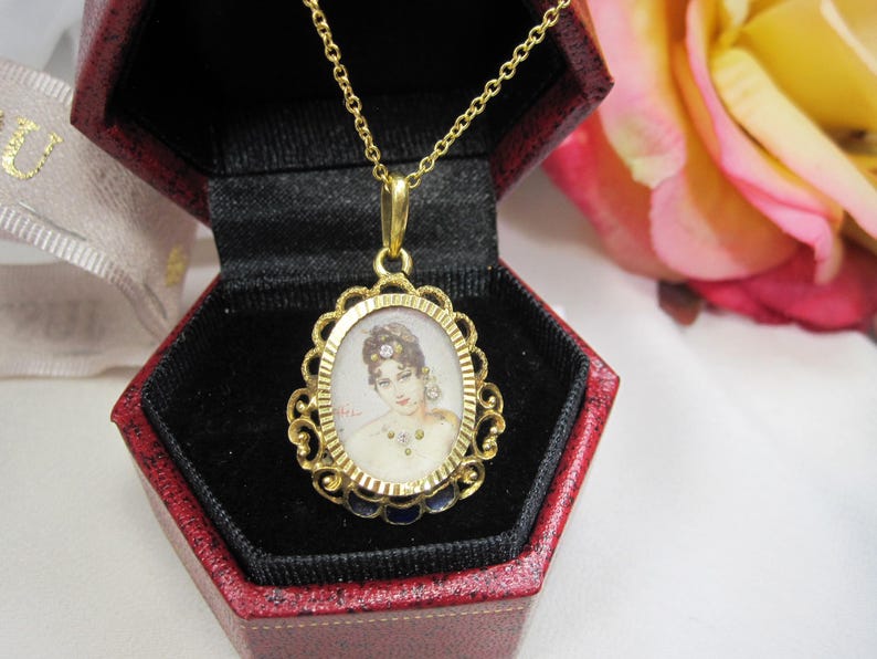 May include: A gold-toned pendant necklace featuring an oval portrait of a woman with a decorative filigree frame. The pendant hangs from a delicate chain and is displayed in a red jewelry box with a black interior.