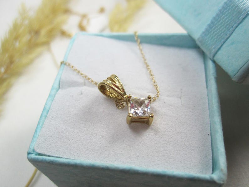May include: A gold chain necklace with a square-shaped pendant featuring a clear cubic zirconia stone.