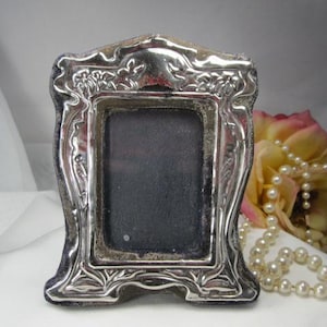 May include: A small, ornate silver picture frame with a rectangular opening. The frame has a detailed floral and scroll design. The background includes a rose and a strand of pearls.