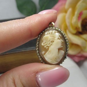 May include: An oval-shaped cameo pendant featuring a carved cream-colored profile of a person facing right. The central carving is set within a metallic frame bordered by a series of small, round, white beads and includes a small loop at the top.