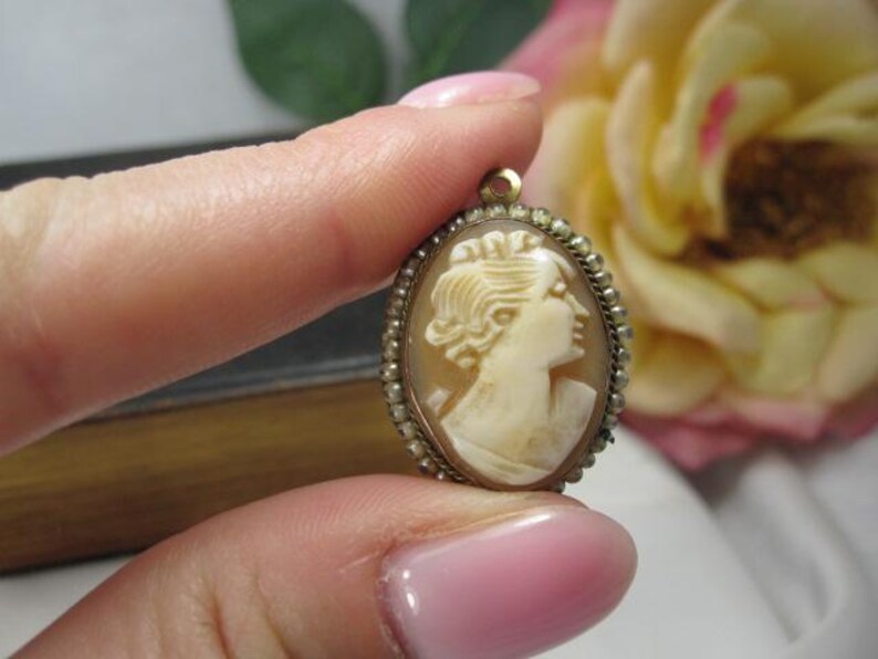 May include: An oval-shaped cameo pendant featuring a carved cream-colored profile of a person facing right. The central carving is set within a metallic frame bordered by a series of small, round, white beads and includes a small loop at the top.