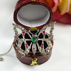 May include: A silver butterfly brooch with clear and red gemstones, centered with a green stone. The brooch is displayed in a red jewelry box with a gold clasp. A silver chain and safety pin are attached.
