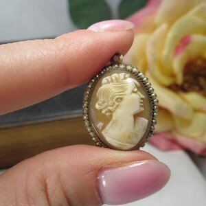May include: An oval-shaped cameo pendant featuring a carved cream-colored profile of a person facing right, set within a metallic frame bordered by a series of small, round, white seed pearls and a small metal loop at the top for attachment.