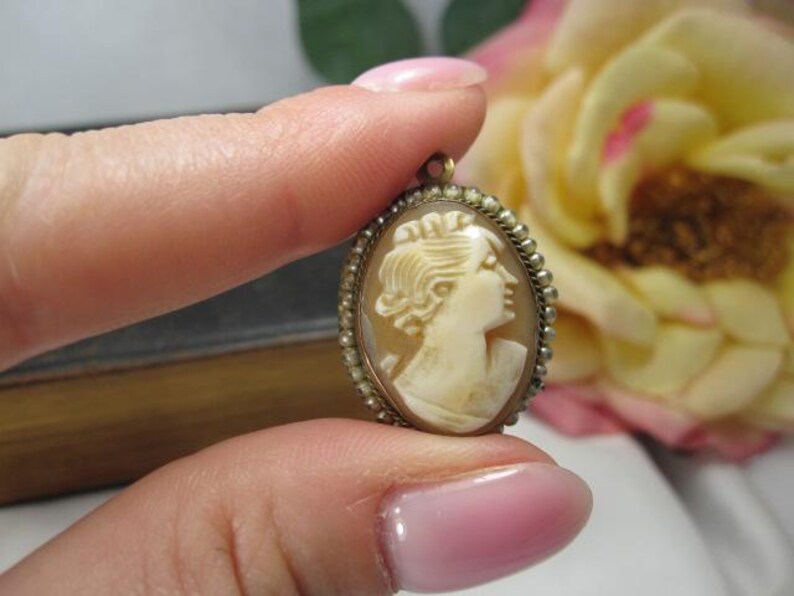 May include: An oval-shaped cameo pendant featuring a carved cream-colored profile of a person facing right, set within a metallic frame bordered by a series of small, round, white seed pearls and a small metal loop at the top for attachment.