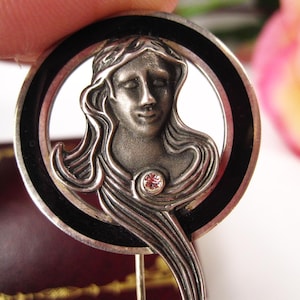 May include: A silver-toned Art Nouveau style pin with a woman's face in a circular frame. The frame has a black enamel border. A small pink gemstone is set below the face. The pin is held by a hand.