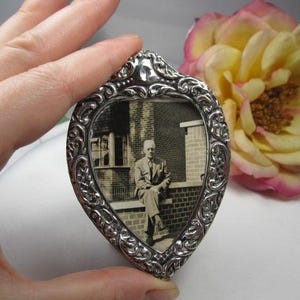 May include: A heart-shaped silver-toned picture frame with an ornate, scrolling design. The frame holds a black and white photograph of a man seated outdoors. A yellow and pink flower is in the background.