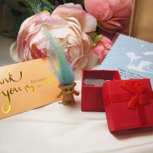 May include: A small, brown troll doll with blue and yellow hair stands near a red gift box with a bow. A peach-colored card with gold lettering that says "Thank you" is also present. The image has a soft, romantic feel with flowers in the background.