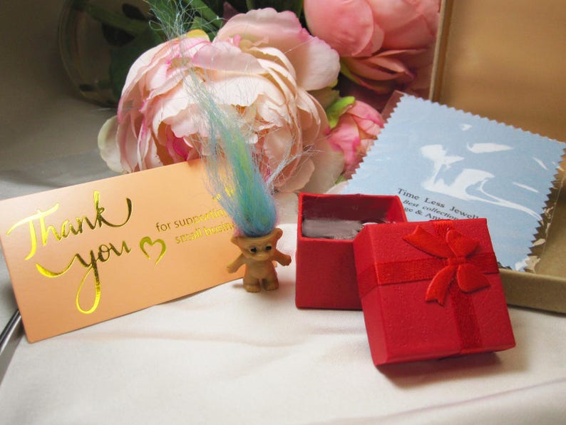 May include: A small, brown troll doll with blue and yellow hair stands near a red gift box with a bow. A peach-colored card with gold lettering that says "Thank you" is also present. The image has a soft, romantic feel with flowers in the background.