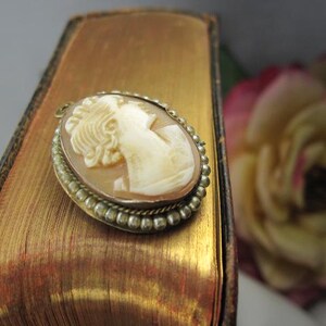 May include: An oval-shaped cameo brooch featuring a carved cream-colored profile of a person against a light brown background. The cameo is set in a metallic frame with a beaded edge and a twisted rope detail, including a small loop at the top.