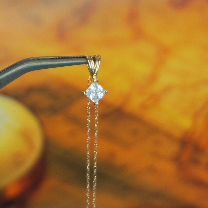 May include: A delicate gold chain necklace with a square-shaped pendant featuring a clear, faceted gemstone.