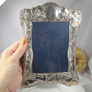 May include: A silver-toned metal picture frame featuring an ornate, embossed floral border with poppy-like flowers at the corners and top center. The frame has a rectangular opening currently filled with a dark blue insert and a smooth, reflective finish.