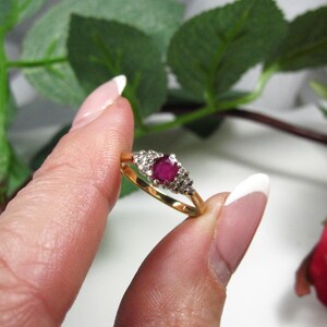 May include: A gold ring featuring a central ruby and surrounding small diamonds. The ring is held by fingers, with green leaves and a red and yellow flower in the background.