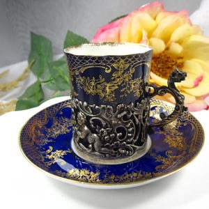 Könnte beinhalten: Ein Vintage-Teebecher- und Untertassen-Set. Die Tasse ist marineblau mit goldenen Blumendesigns und einem silbernen Filigranhalter. Die Untertasse ist ebenfalls marineblau mit goldenen Akzenten. Der Griff hat ein Drachendesign.