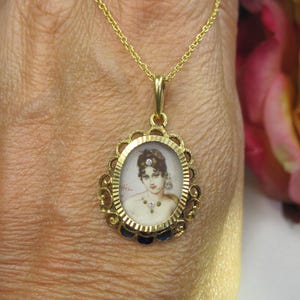 May include: Gold pendant necklace featuring an oval portrait of a woman. The pendant has a decorative gold frame with a scalloped edge and small blue accents. The chain is a delicate gold link.