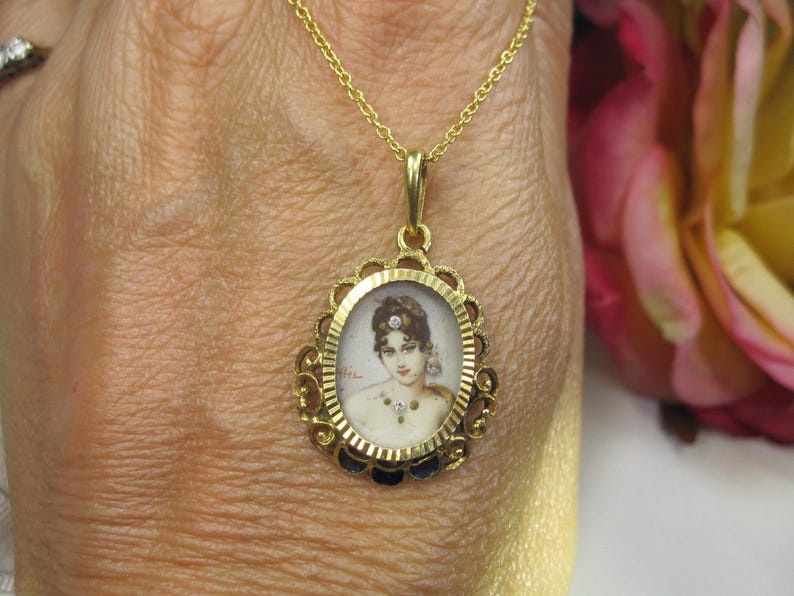 May include: Gold pendant necklace featuring an oval portrait of a woman. The pendant has a decorative gold frame with a scalloped edge and small blue accents. The chain is a delicate gold link.