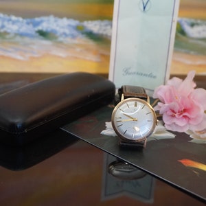 Op de afbeelding: Een vintage goudkleurig polshorloge met een lichtgekleurde wijzerplaat en een donker leren bandje. Het horloge wordt gepresenteerd met een zwarte etui, een roze bloem en een kaart met de tekst "Guarantee". De wijzerplaat van het horloge heeft goudkleurige wijzers en markeringen.