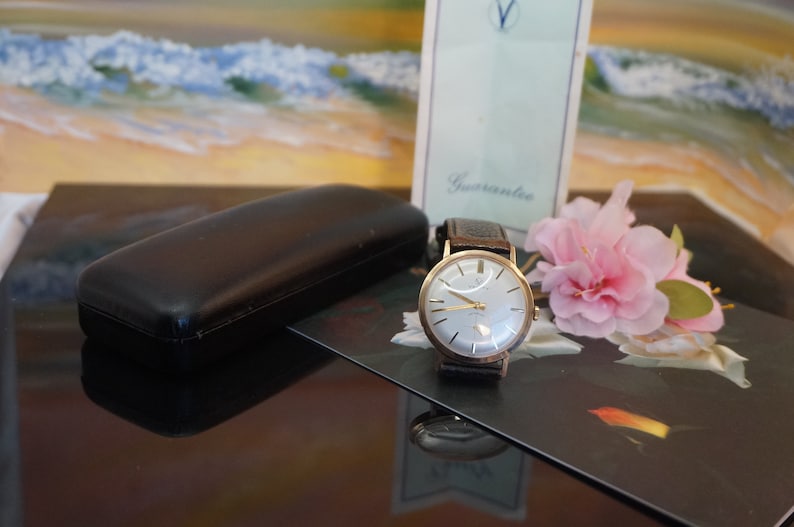 Op de afbeelding: Een vintage goudkleurig polshorloge met een lichtgekleurde wijzerplaat en een donker leren bandje. Het horloge wordt gepresenteerd met een zwarte etui, een roze bloem en een kaart met de tekst "Guarantee". De wijzerplaat van het horloge heeft goudkleurige wijzers en markeringen.