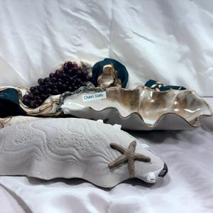 May include: Two ceramic serving dishes are displayed. One is a clam shell shape with a starfish attached. The other is a three-sectioned dish with a wavy rim. A bunch of grapes and a patterned scarf are also in the image. The three-sectioned dish has the text "Oven Safe".