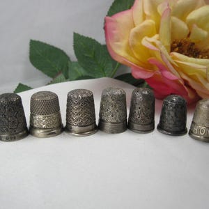 May include: A collection of seven metallic thimbles arranged in a row, each featuring distinct embossed patterns including floral motifs, geometric grids, and one thimble with the word HOVIS clearly engraved around the base rim.