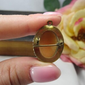 May include: An oval-shaped brooch featuring a smooth, light peach-colored center inlay set within a tarnished brass-toned metal frame. The back of the piece includes a horizontal pin mechanism and a small circular loop at the top for hanging.