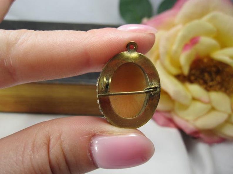 May include: An oval-shaped brooch featuring a smooth, light peach-colored center inlay set within a tarnished brass-toned metal frame. The back of the piece includes a horizontal pin mechanism and a small circular loop at the top for hanging.
