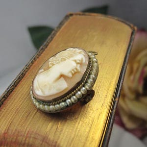 May include: An oval-shaped cameo brooch featuring a carved profile of a person in cream-colored material against a light brown background. The brooch is framed by a border of small, round, white pearls set in a metallic, textured gold-tone setting.