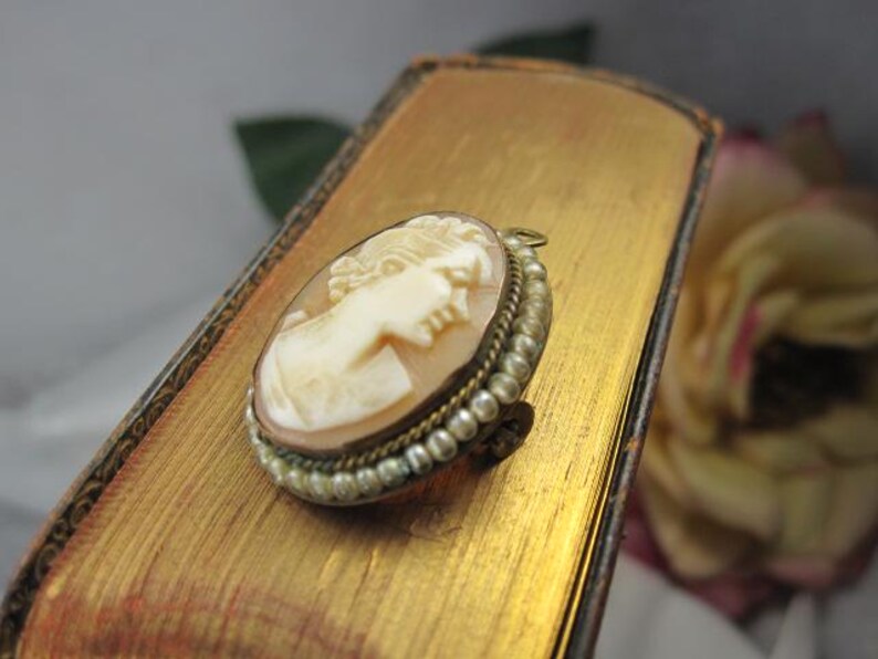 May include: An oval-shaped cameo brooch featuring a carved profile of a person in cream-colored material against a light brown background. The brooch is framed by a border of small, round, white pearls set in a metallic, textured gold-tone setting.