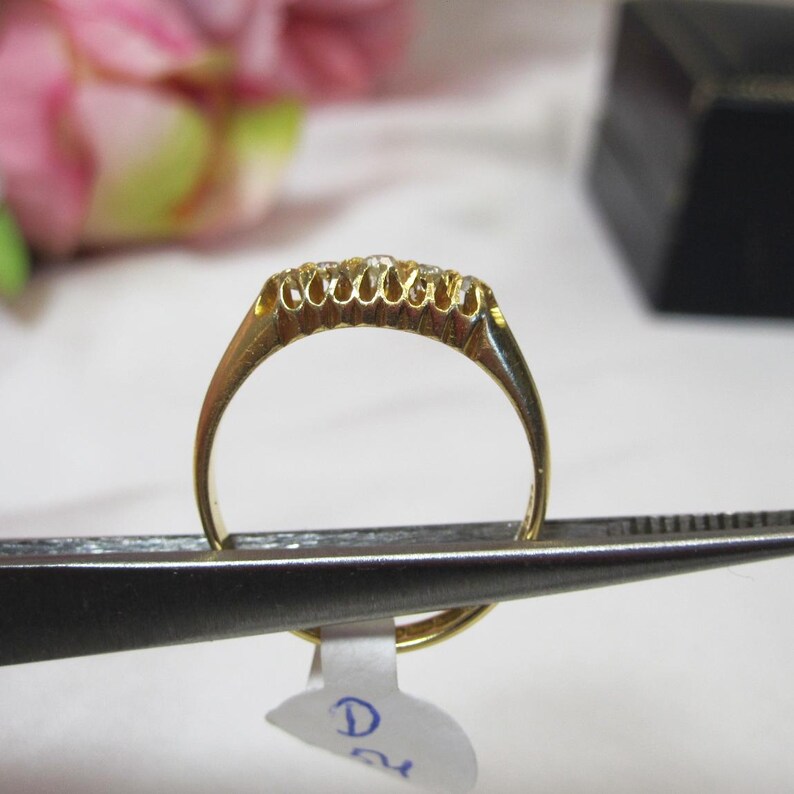 May include: A gold band ring with a row of small diamonds set in the band. The ring is held by a pair of tweezers.
