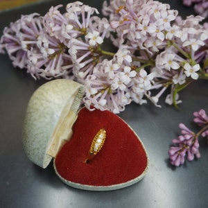 May include: A gold ring with small diamonds set in a heart-shaped ring box lined with red velvet. The box is surrounded by lilac flowers.