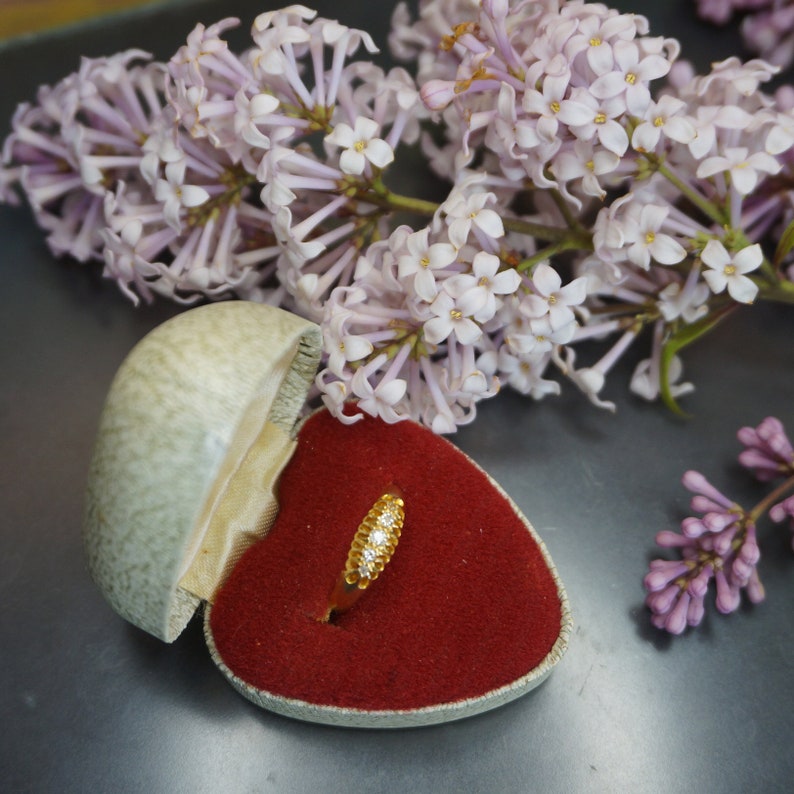 May include: A gold ring with small diamonds set in a heart-shaped ring box lined with red velvet. The box is surrounded by lilac flowers.