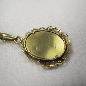 May include: A gold-colored pendant necklace with an oval-shaped pendant. The pendant has a decorative, openwork border and a textured surface. The chain is delicate and also gold-colored. The necklace is displayed on a white surface.