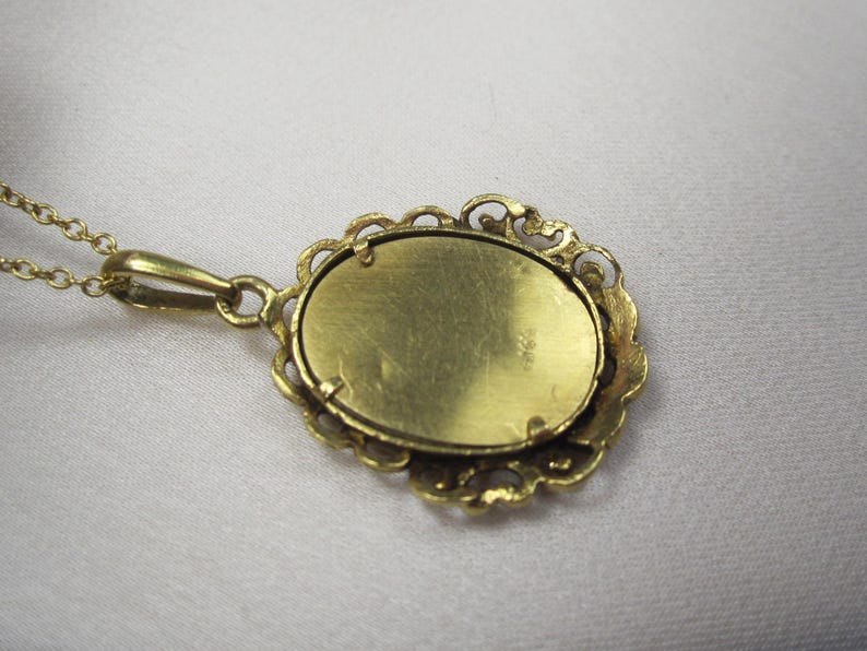 May include: A gold-colored pendant necklace with an oval-shaped pendant. The pendant has a decorative, openwork border and a textured surface. The chain is delicate and also gold-colored. The necklace is displayed on a white surface.