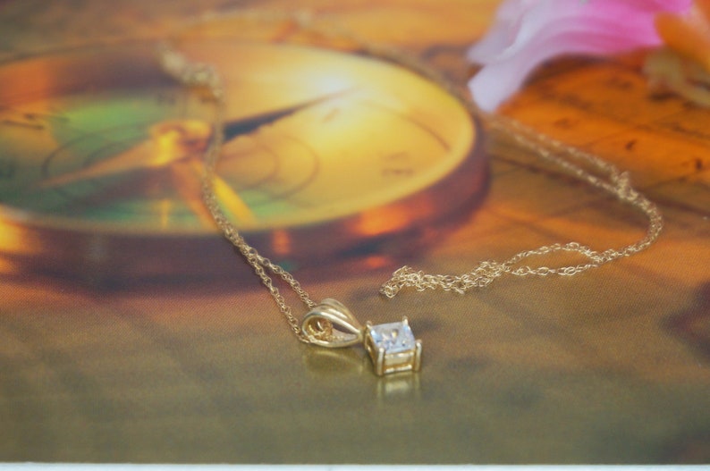 May include: A gold chain necklace with a square-shaped pendant featuring a clear gemstone.