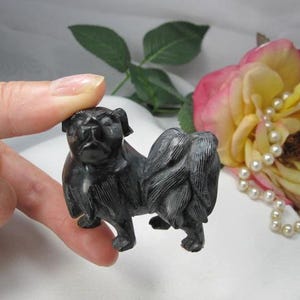 May include: A small, black, carved stone figurine of a dog with a flat face, prominent eyes, and a bushy, curled tail. The sculpture features detailed, etched lines representing fur texture across its body, standing on four small, distinct paws.