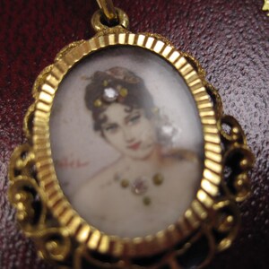 May include: A gold-toned oval pendant with a miniature portrait of a woman. The portrait features a woman with dark hair, red lips, and a jeweled necklace. The pendant has an ornate, filigree-style frame.