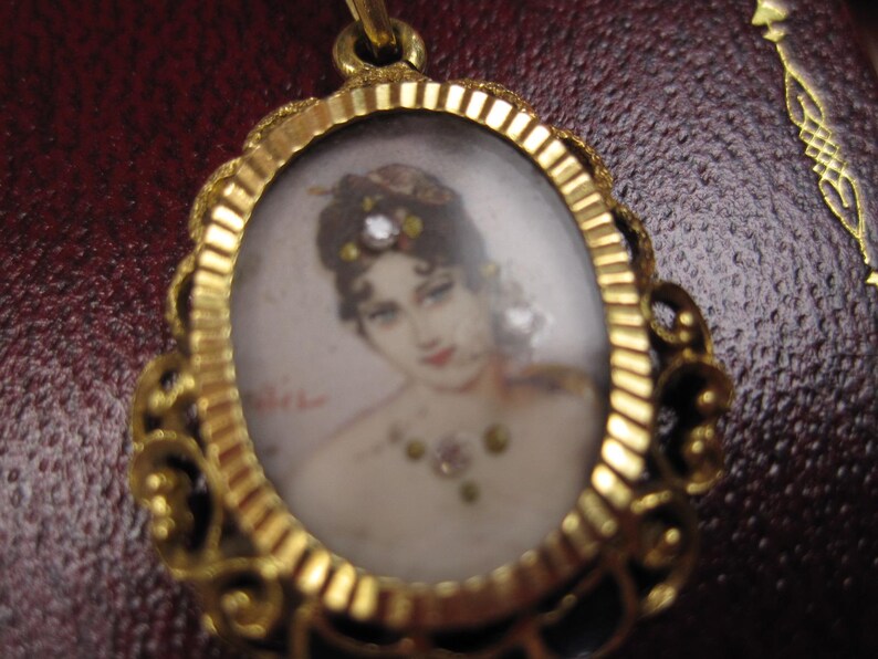 May include: A gold-toned oval pendant with a miniature portrait of a woman. The portrait features a woman with dark hair, red lips, and a jeweled necklace. The pendant has an ornate, filigree-style frame.