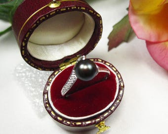 Puede incluir: Un anillo de perla negra con una banda de plata y una fila de pequeñas piedras transparentes. El anillo se exhibe en un joyero redondo, burdeos, abierto, con ribete dorado e interior color crema.