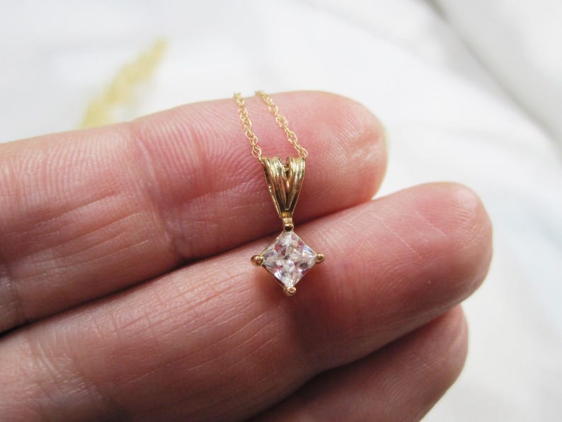 May include: A gold chain necklace with a square-shaped pendant featuring a clear cubic zirconia stone.
