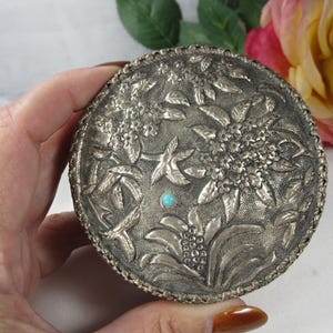 Peut inclure: Une boîte ronde en métal argenté présentant un motif floral en relief complexe sur le couvercle. La surface affiche des feuilles et des pétales détaillés avec une petite pierre ronde incrustée de couleur bleu turquoise au centre.