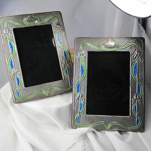 May include: Two rectangular metal picture frames featuring a textured silver-tone finish with decorative green and blue enamel accents. The frames have a central opening for a photograph and a repeating pattern of curved lines and floral motifs.