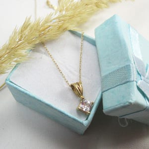 May include: A gold chain necklace with a square-shaped clear crystal pendant. The necklace is in a light blue gift box with a white ribbon.