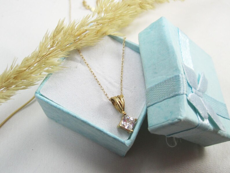 May include: A gold chain necklace with a square-shaped clear crystal pendant. The necklace is in a light blue gift box with a white ribbon.