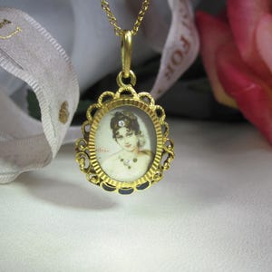 May include: A gold pendant necklace featuring an oval portrait of a woman. The portrait is set within a decorative, scalloped frame. The necklace is on a white surface, with a ribbon and a pink rose in the background.