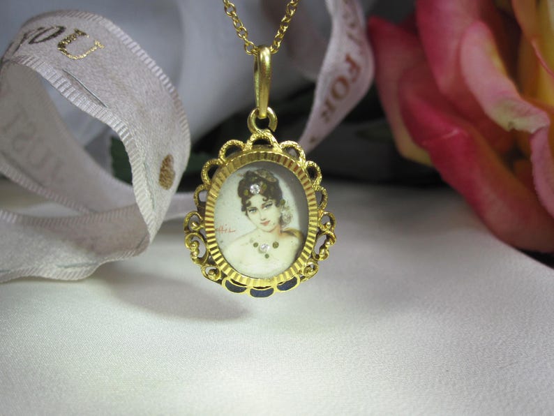 May include: A gold pendant necklace featuring an oval portrait of a woman. The portrait is set within a decorative, scalloped frame. The necklace is on a white surface, with a ribbon and a pink rose in the background.