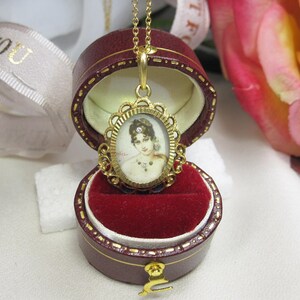 May include: A gold-toned pendant necklace with an oval portrait of a woman, set in an ornate frame. The pendant is displayed in a burgundy jewelry box with a red velvet interior. A white ribbon with gold text is in the background.