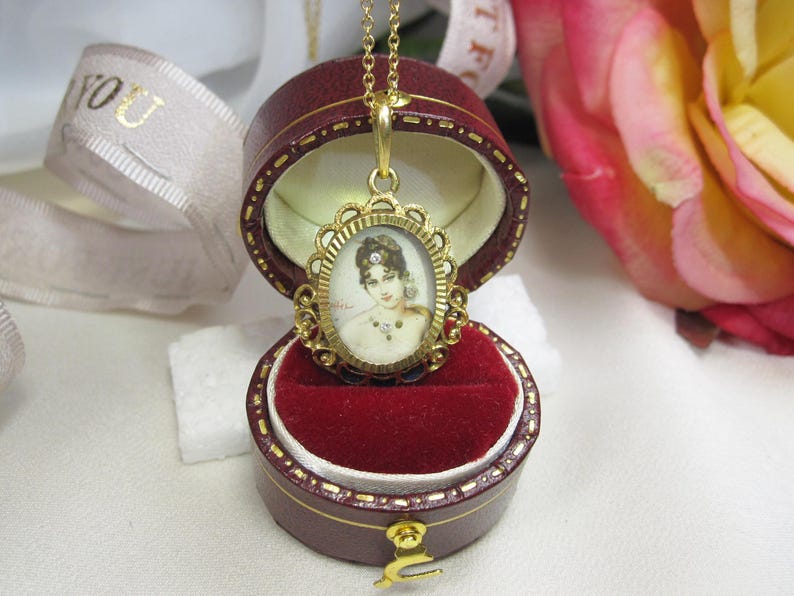 May include: A gold-toned pendant necklace with an oval portrait of a woman, set in an ornate frame. The pendant is displayed in a burgundy jewelry box with a red velvet interior. A white ribbon with gold text is in the background.