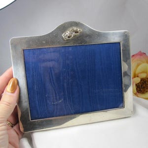 May include: A rectangular silver-toned picture frame with a smooth, polished finish. The frame features a central arched top detail with a small, decorative metallic emblem. The interior display area is filled with a solid, deep blue fabric insert.