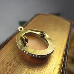 May include: An oval-shaped brooch featuring a central peach-colored cameo inlay surrounded by a border of small white pearls set in a gold-toned metal frame. The back of the brooch displays a metal pin mechanism and a small loop for hanging.