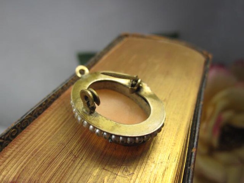 May include: An oval-shaped brooch featuring a central peach-colored cameo inlay surrounded by a border of small white pearls set in a gold-toned metal frame. The back of the brooch displays a metal pin mechanism and a small loop for hanging.